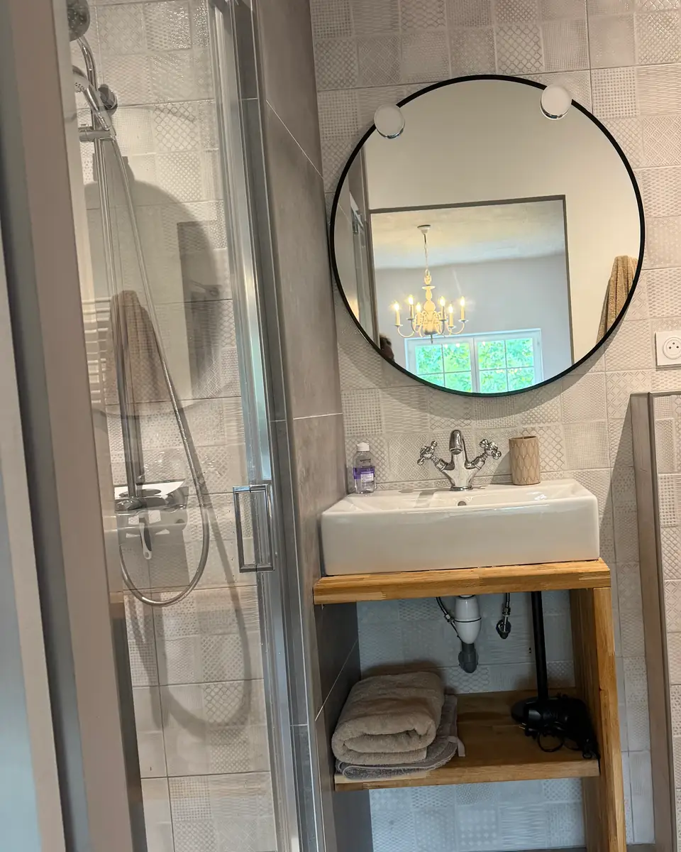 Een badkamer met grijze patroontegels, een glazen douche en een ronde zwarte spiegel.