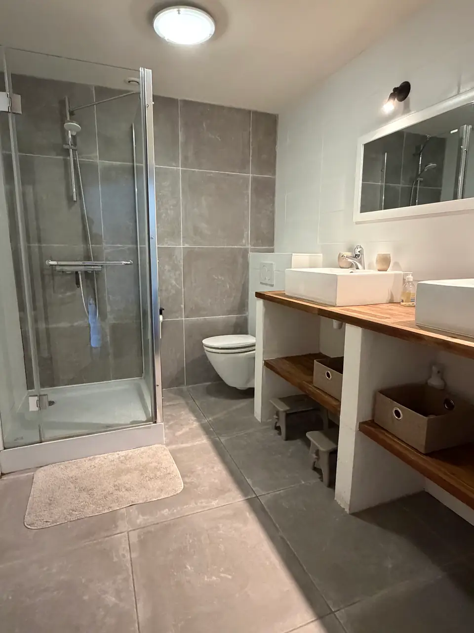 Een badkamer met grote grijze tegels, een glazen douche en twee wastafels op een houten blad.