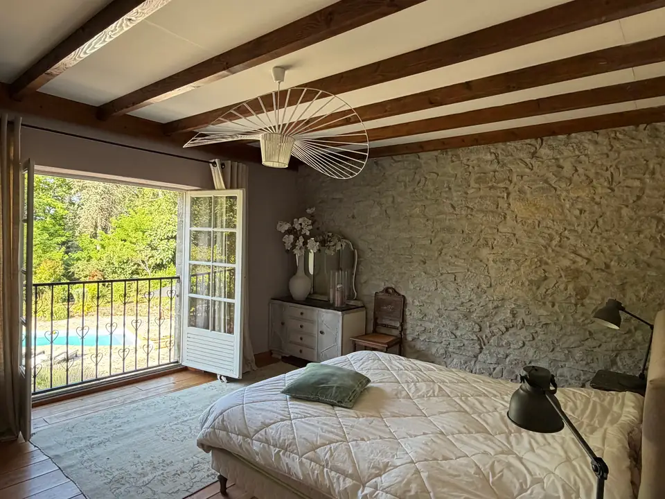 Een slaapkamer met een stenen muur en balken; de openslaande deuren komen uit op een ijzeren balkon boven het zwembad.