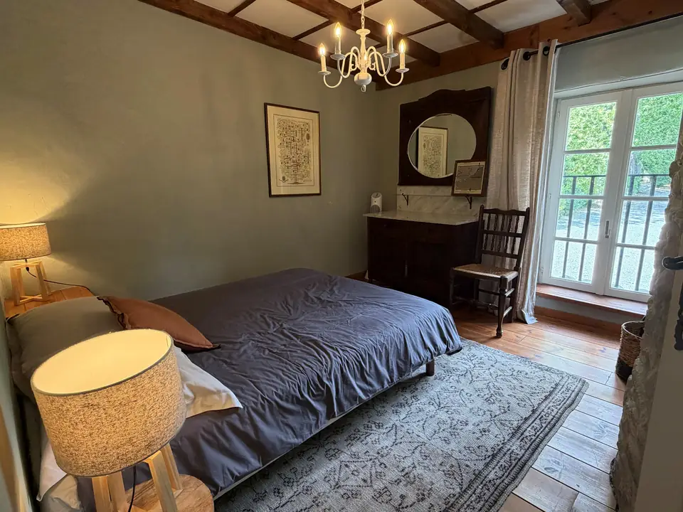 Een slaapkamer met grijsgroene muren, een kroonluchter, een lampetkast met spiegel en openslaande deuren.