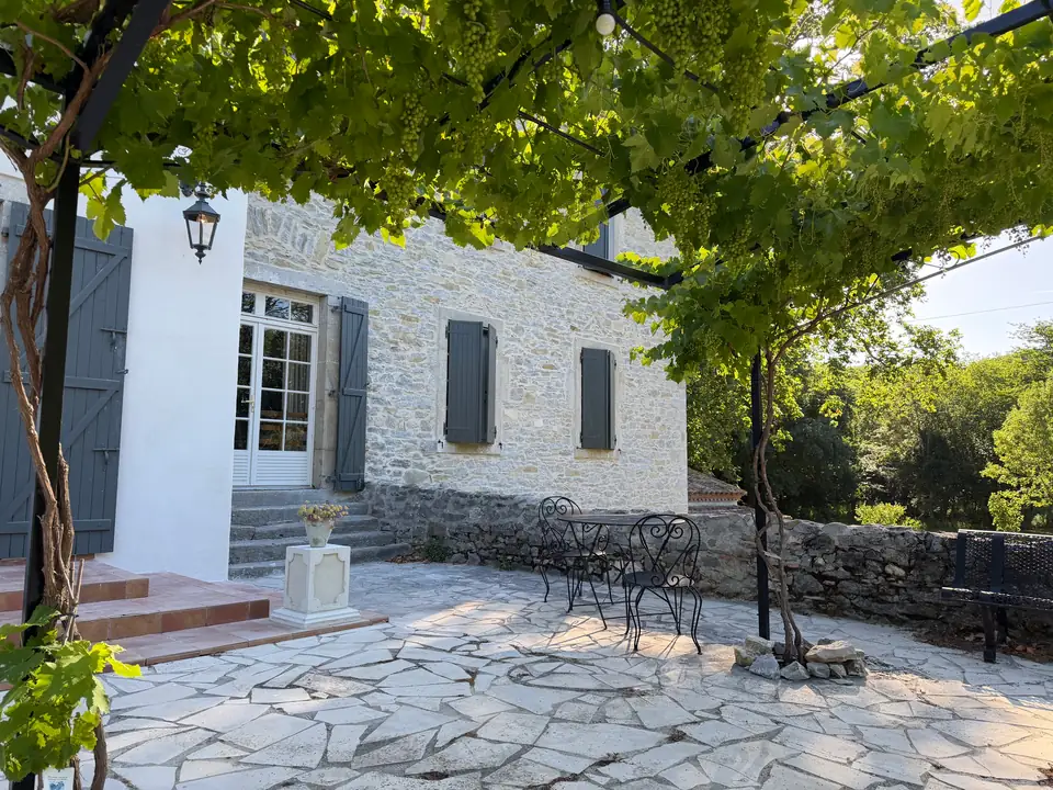 Onder de pergola hangen de druiven; erachter de witte vleugel en het stenen huis.